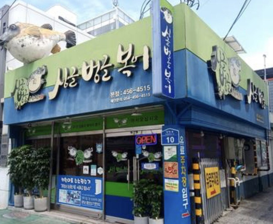파랑·연두 모퉁이 가게 외관 — 지붕 위 복어 조형, '싱글벙글 복어' 간판(정면·옆면·세로 간판), 전화번호 '456-4515', 번지판 '10', 'OPEN' 네온, 화분