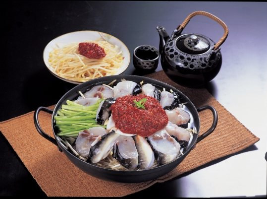 검은 양손 냄비에 껍질째 썬 복어 살과 미나리를 둘러 담고 가운데에 붉은 양념을 올린 끓이기 전 모습 — 뒤에 붉은 양념을 올린 콩나물무침·검은 찻주전자·찻잔